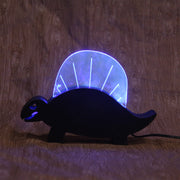 Wooden sculpture of a black dinosaur, flat mostly and emphasizing its silhouette.  Atop its back is a rounded plastic sheet with a line of lights, illuminating it and the interior of its mouth.