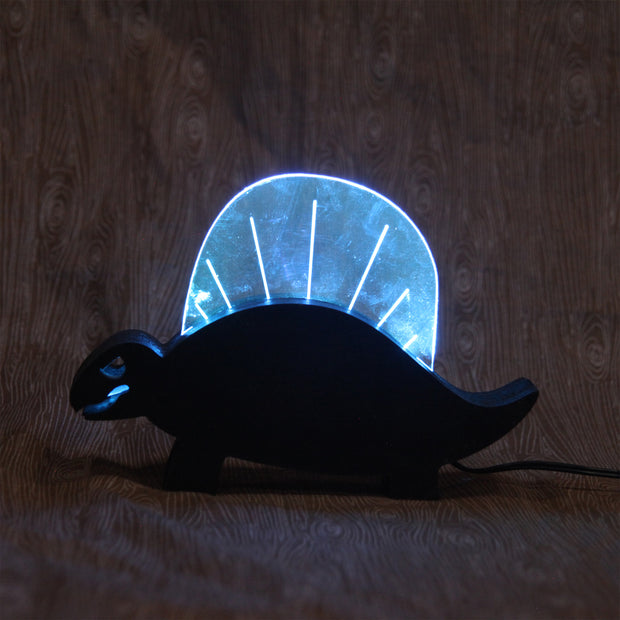 Wooden sculpture of a black dinosaur, flat mostly and emphasizing its silhouette.  Atop its back is a rounded plastic sheet with a line of lights, illuminating it and the interior of its mouth.