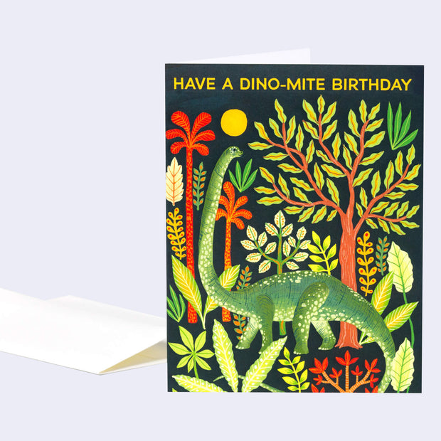 Black greeting card with an illustration of a green brachiosaurus amongst greenery and a yellow sun. Text above reads "Have a Dino-Mite Birthday"