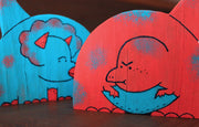 2 die cut painted wooden dinosaur sculptures, one a blue cartoon triceratops and the other a red cartoon simplistic dinosaur. 