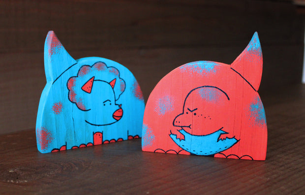 2 die cut painted wooden dinosaur sculptures, one a blue cartoon triceratops and the other a red cartoon simplistic dinosaur. 