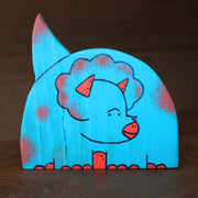 Die cut painted wooden sculpture of a blue chubby cartoon triceratops dinosaur. It is blue with red patchy spots and accent colorings.