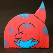 Painted die cut wooden sculpture of a red cartoon dinosaur with a rounded head and pointed tail. It has a cute, angry expression and has blue patchy spots on its body and a blue belly.