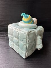 Ceramic sculpture of Snoopy wearing a puffy teal colored jacket and blue and yellow striped beanie. His body is shaped like a cube.