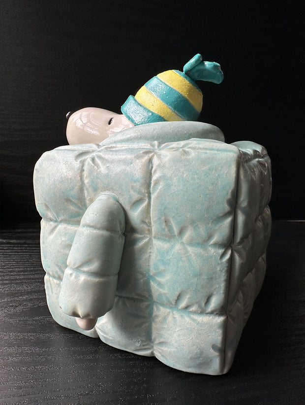 Ceramic sculpture of Snoopy wearing a puffy teal colored jacket and blue and yellow striped beanie. His body is shaped like a cube.