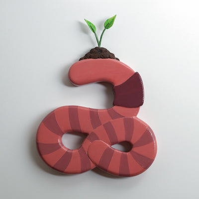 Die cut flat painted sculpture of a pink earthworm, with a pile of dirt atop its head and 2 sprouts growing out of it.