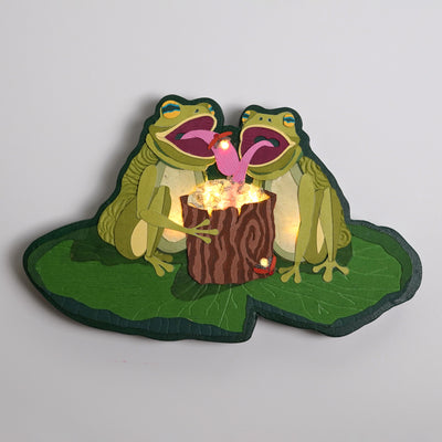 Die cut sculpture of 2 frogs sitting on a large lily pad and extending their tongues out into a hollowed out log holding many lit up lightening bugs. They each have their tongues out, catching the bugs.