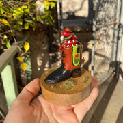 Sculpture of a red snake with black and white stripes, coiled up inside of a brown and black cowboy boot with a spur. Cowboy boot has a design element of a pink tulip with yellow stars on green background on each side of the boot. Snake wears a small cowboy hat and bandana. 