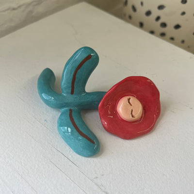 Shiny ceramic sculpture of a red flower with a simplistic green stem and leaves, resting on the ground with a pair of closed eyes.