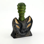 Whittled wooden sculpture of the bust of a bat/wolf creature, black with wooden accents. Coming out of its head is a green bumpy succulent cactus.