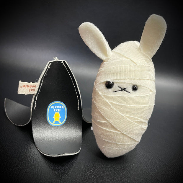 Small plush rabbit, with different sized eyes and white linen wrappings around its body like a mummy. Next to it is an upside down black banana peel.