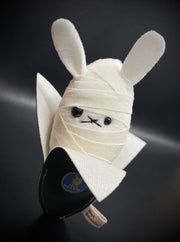 Small plush rabbit, with different sized eyes and white linen wrappings around its body like a mummy. It sits inside of a black banana peel.