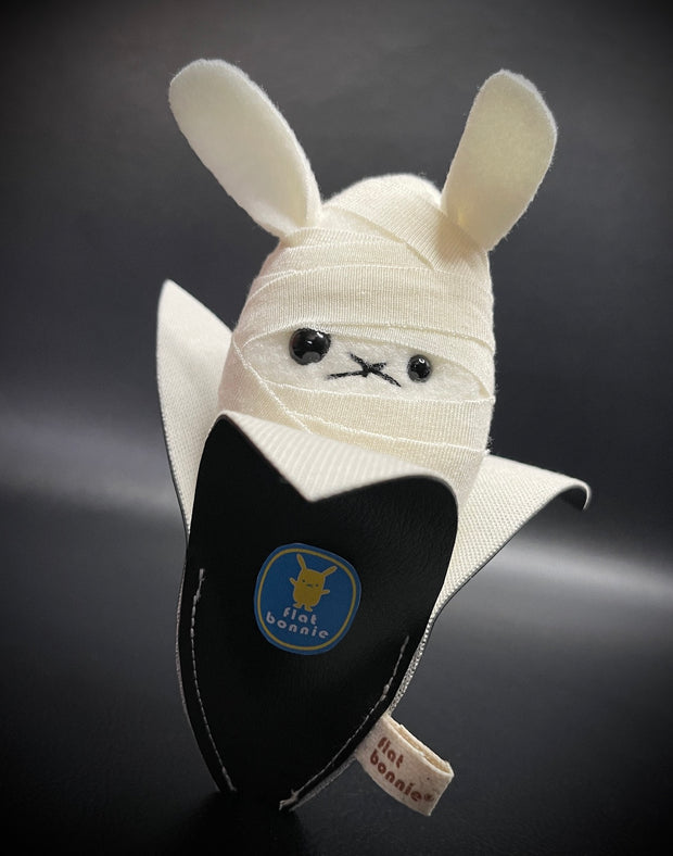 Small plush rabbit, with different sized eyes and white linen wrappings around its body like a mummy. It sits inside of a black banana peel.