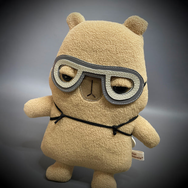 Plush sculpture of a cartoon capybara with a large head and flat form. It has an apathetic expression and wears a pair of pleather flying goggles.