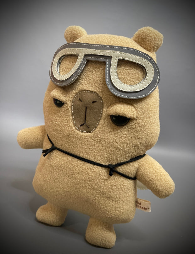 Plush sculpture of a cartoon capybara with a large head and flat form. It has an apathetic expression and wears a pair of pleather flying goggles.