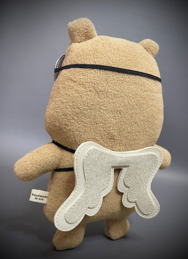 Plush sculpture of a cartoon capybara with a large head and flat form. It has an apathetic expression and wears a pair of pleather flying goggles and a pair of wings strapped to its back.