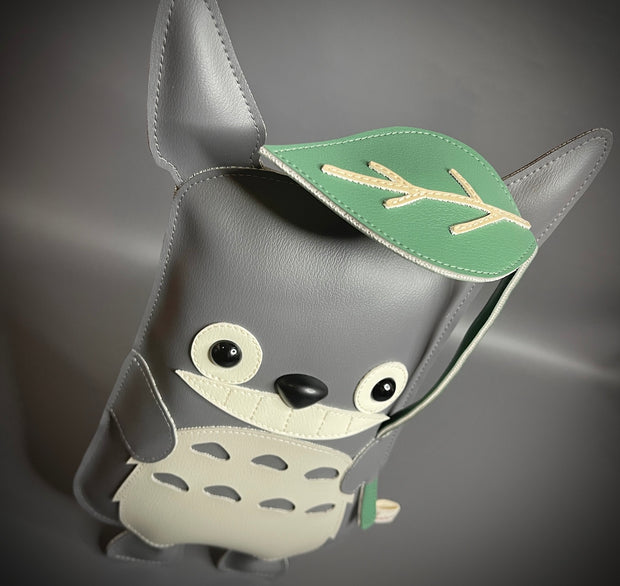 Vinyl plush sculpture of a Totoro inspired character, depicted in a flat style with small round eyes and legs. It has a wide eyed smile and holds a leaf umbrella overhead. 