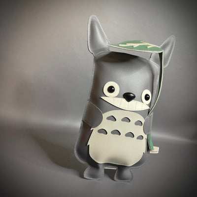 Vinyl plush sculpture of a Totoro inspired character, depicted in a flat style with small round eyes and legs. It has a wide eyed smile and holds a leaf umbrella overhead. 