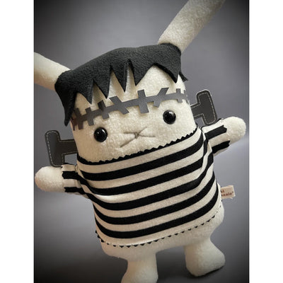 White bunny plush doll, flat in style, dressed as Frankenstein with a black and white striped shirt and stitches on its forehead and bolts coming out of its neck.