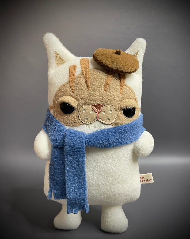 Flat plush doll of a cream colored cat with brown face and stripes atop his head. It wears a small brown beret cap and a blue scarf wrapped around its neck.