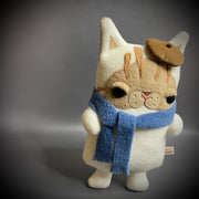 Flat plush doll of a cream colored cat with brown face and stripes atop his head. It wears a small brown beret cap and a blue scarf wrapped around its neck.