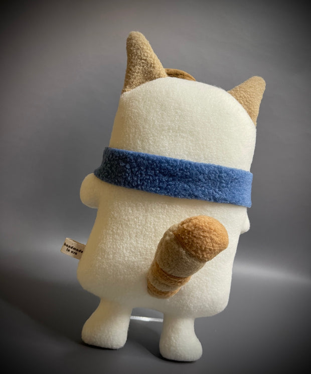 Flat plush doll of a cream colored cat with brown face and stripes atop his head. It wears a small brown beret cap and a blue scarf wrapped around its neck.