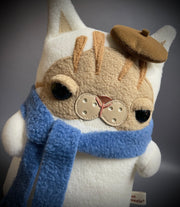 Flat plush doll of a cream colored cat with brown face and stripes atop his head. It wears a small brown beret cap and a blue scarf wrapped around its neck.