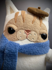 Flat plush doll of a cream colored cat with brown face and stripes atop his head. It wears a small brown beret cap and a blue scarf wrapped around its neck.