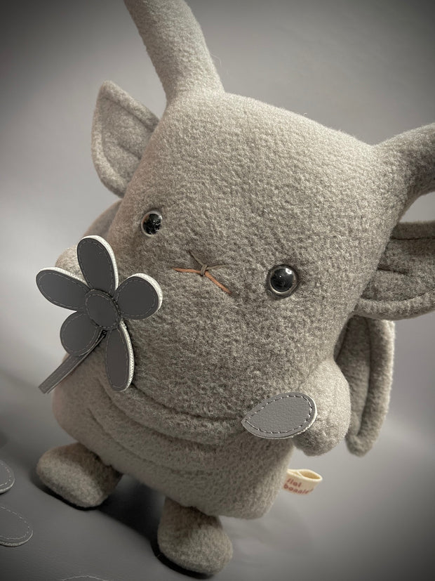 Plush doll of a bunny with skinny ears and a flat body, made to look like a gargoyle. It holds a pleather flower and plucks a petal from it.