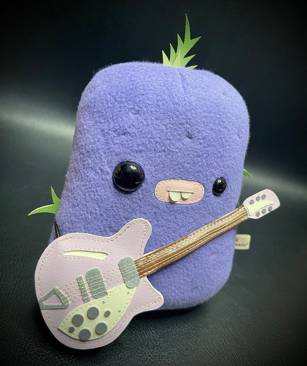 Flat plush sculpture of an ube, a purple root vegetable. Plush has different sized eyes and a bucktooth mouth. It holds a light purple electric guitar.