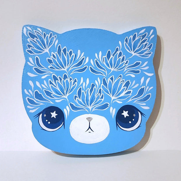 Die cut wooden panel of a cat's head, painted blue with an intricate floral design on its forehead and eyebrows. It has large anime style eyes, dark blue with white stars and a small mouth.