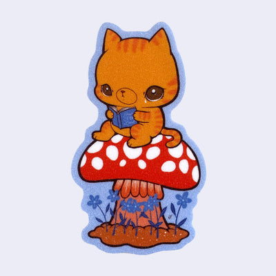 Die cut sticker with a glittery surface of an orange cat sitting atop of a red mushroom and crying while reading a book.