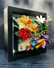 Ceramic sculpture in an open shadow box frame featuring many flowers, alike to the ones from Alice in Wonderland. A small blonde girl stands off to the side and looks onward with her back facing the viewer.