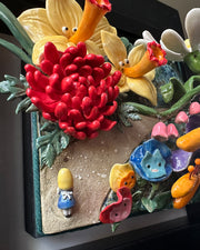 Ceramic sculpture in an open shadow box frame featuring many flowers, alike to the ones from Alice in Wonderland. A small blonde girl stands off to the side and looks onward with her back facing the viewer.