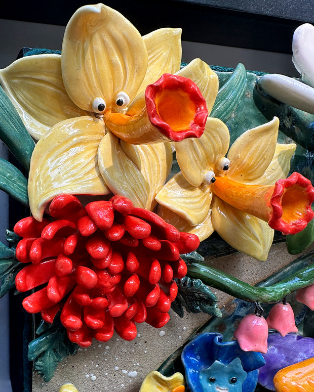 Ceramic sculpture in an open shadow box frame featuring many flowers, alike to the ones from Alice in Wonderland. A small blonde girl stands off to the side and looks onward with her back facing the viewer.