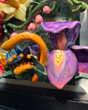 Ceramic sculpture in an open shadow box frame featuring many flowers, alike to the ones from Alice in Wonderland. A small blonde girl stands off to the side and looks onward with her back facing the viewer.