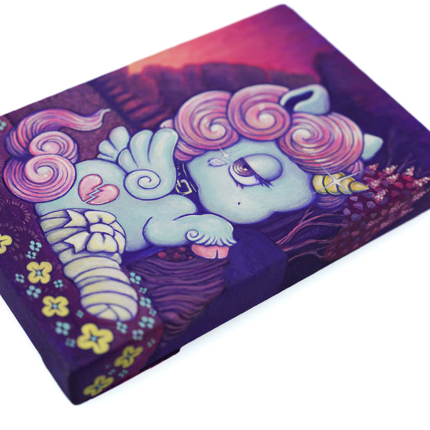 Painting of a small, cute blue cartoon unicorn sitting on the ground with a bandaged leg. It has a pink curly mane and a sparkly horn. Background is a red and purple dark sunset.