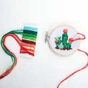 Cross stitch embroidery in a bamboo circle of a set of cacti plants, green with red and orange blossoms. Colored thread is next to the hoop.