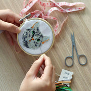 Someone working on an embroidery of tabby cat's head with green eyes on a desk with fabric measuring tape, small scissors and colorful threads.