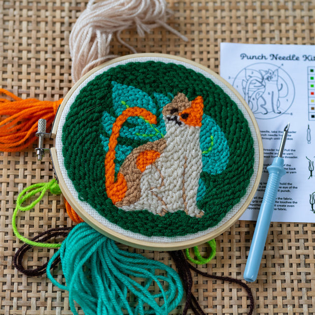 All contents of a Punch Needle Kit with the design of an orange cat with a large Monstera style leaf behind it on a dark green background. Includes various colored thread, punch needle, instructions, an embroidery hoop.