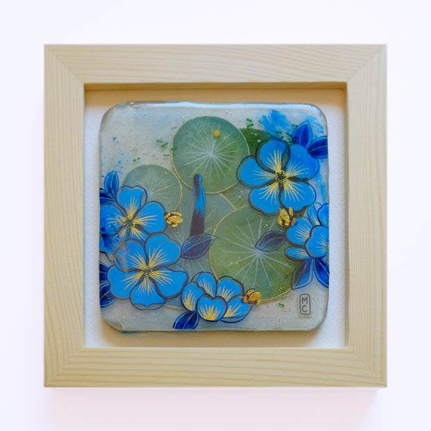 Resin coated square featuring green lilypads and blue and gold flowers. A slender blue woman stands in the middle and looks off, with gold bees buzzing around. Piece is in glassless wooden frame.