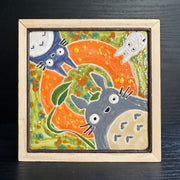 Painted ceramic tile with bright green and orange coloring with 3 characters surrounding the exterior. One is Totoro with a leaf umbrella, the other is a dark blue smaller Totoro and the last is a small white Totoro.