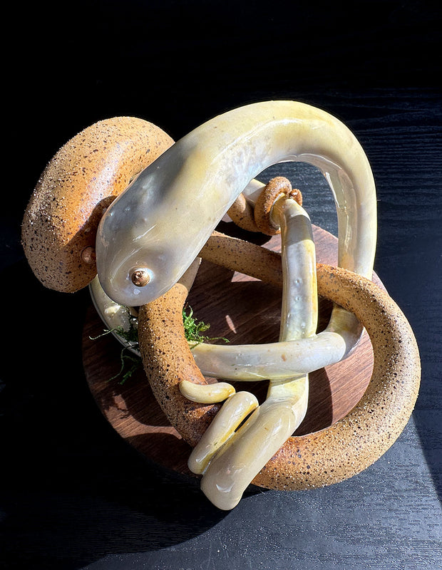 Ceramic sculpture of 2 brown snakes, intertwined with one another. One is matte brown like sand and the other is a glossy yellow. They both have golden eyes and their faces pressed together.