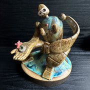 Ceramic sculpture of a brown robot with a small head, and large arms and body. It sits atop of a blue glazed mound and extends out one of its hands. It holds a dirt clod with a pink flower growing out of it.