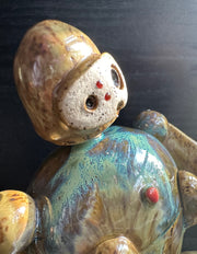 Ceramic sculpture of a brown robot with a small head, and large arms and body. It sits atop of a blue glazed mound and extends out one of its hands. It holds a dirt clod with a pink flower growing out of it.