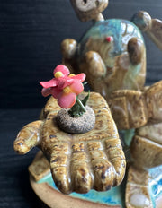 Ceramic sculpture of a brown robot with a small head, and large arms and body. It sits atop of a blue glazed mound and extends out one of its hands. It holds a dirt clod with a pink flower growing out of it.