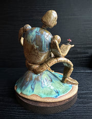 Ceramic sculpture of a brown robot with a small head, and large arms and body. It sits atop of a blue glazed mound and extends out one of its hands. It holds a dirt clod with a pink flower growing out of it.