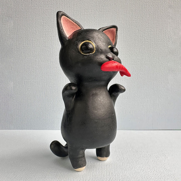 Ceramic sculpture of a black cat with large shiny eyes and pointed ears with pink interiors. It stands on its hind legs and has its paws up in the air and holds a red fish in its mouth.