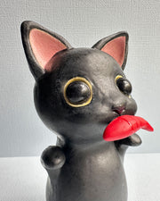 Ceramic sculpture of a black cat with large shiny eyes and pointed ears with pink interiors. It stands on its hind legs and has its paws up in the air and holds a red fish in its mouth.