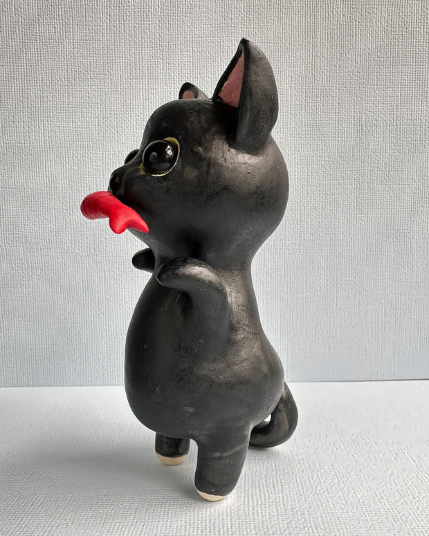 Ceramic sculpture of a black cat with large shiny eyes and pointed ears with pink interiors. It stands on its hind legs and has its paws up in the air and holds a red fish in its mouth.